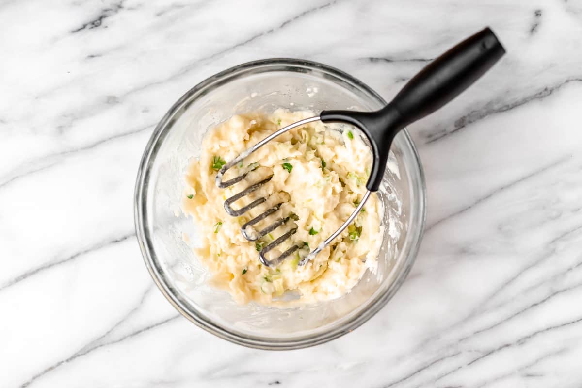 Mashed potatoes with scallions in a glass bowl with a potato masher.