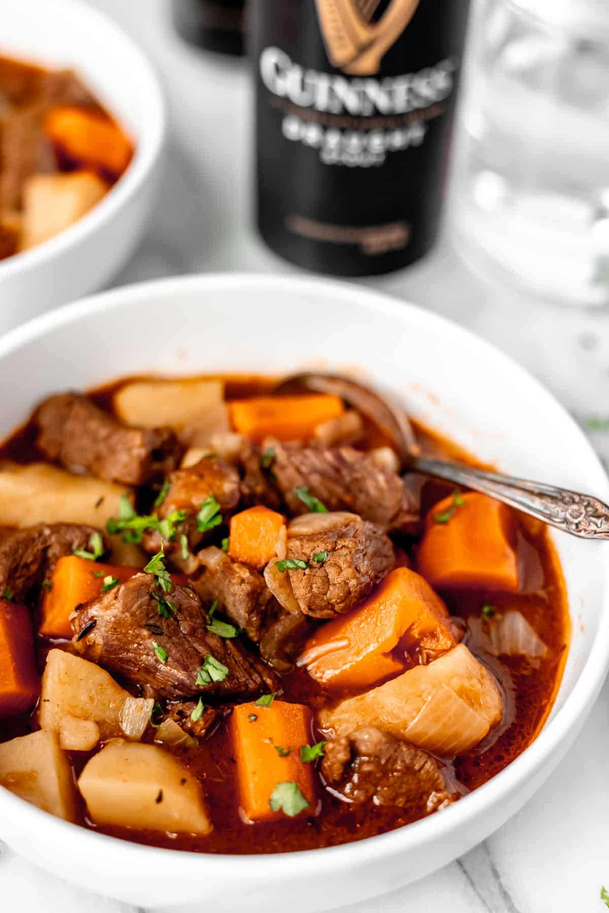 Close up of a bowl of Instant Pot Guinness Beef Stew in a white bowl with a spoon in it and a second bowl and can of Guinness in the background.