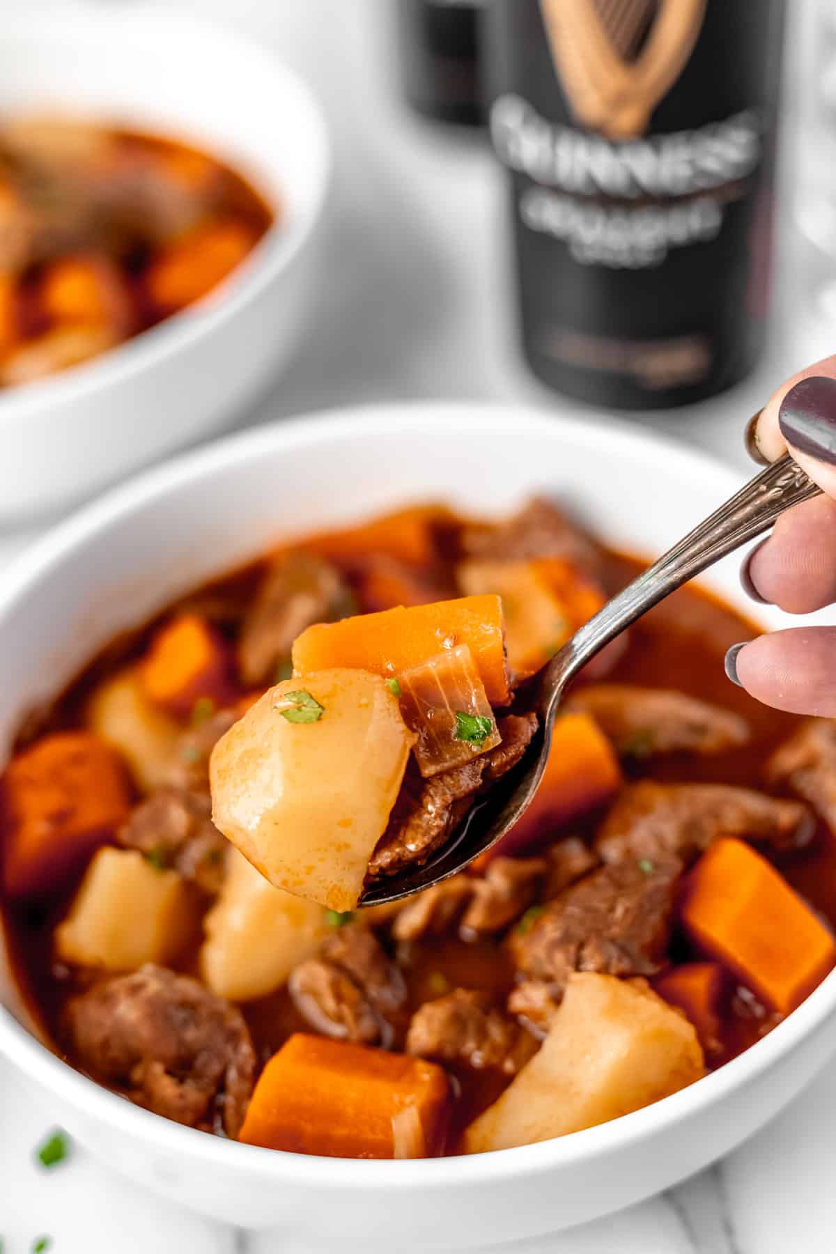 A spoonful of Guinness beef stew being held up in front of the bowl with a can of Guinness in the background.