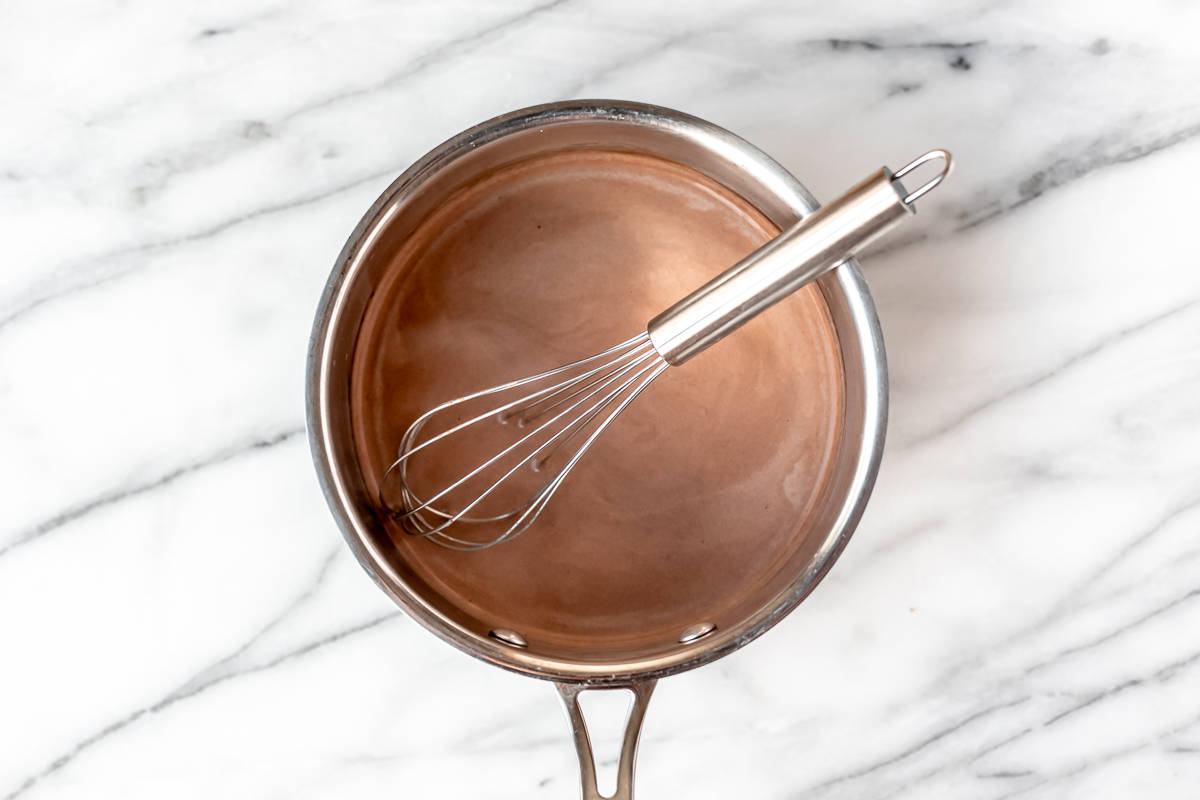 A silver pot filled with milk and cocoa powder with a silver whisk in it over a marble background.