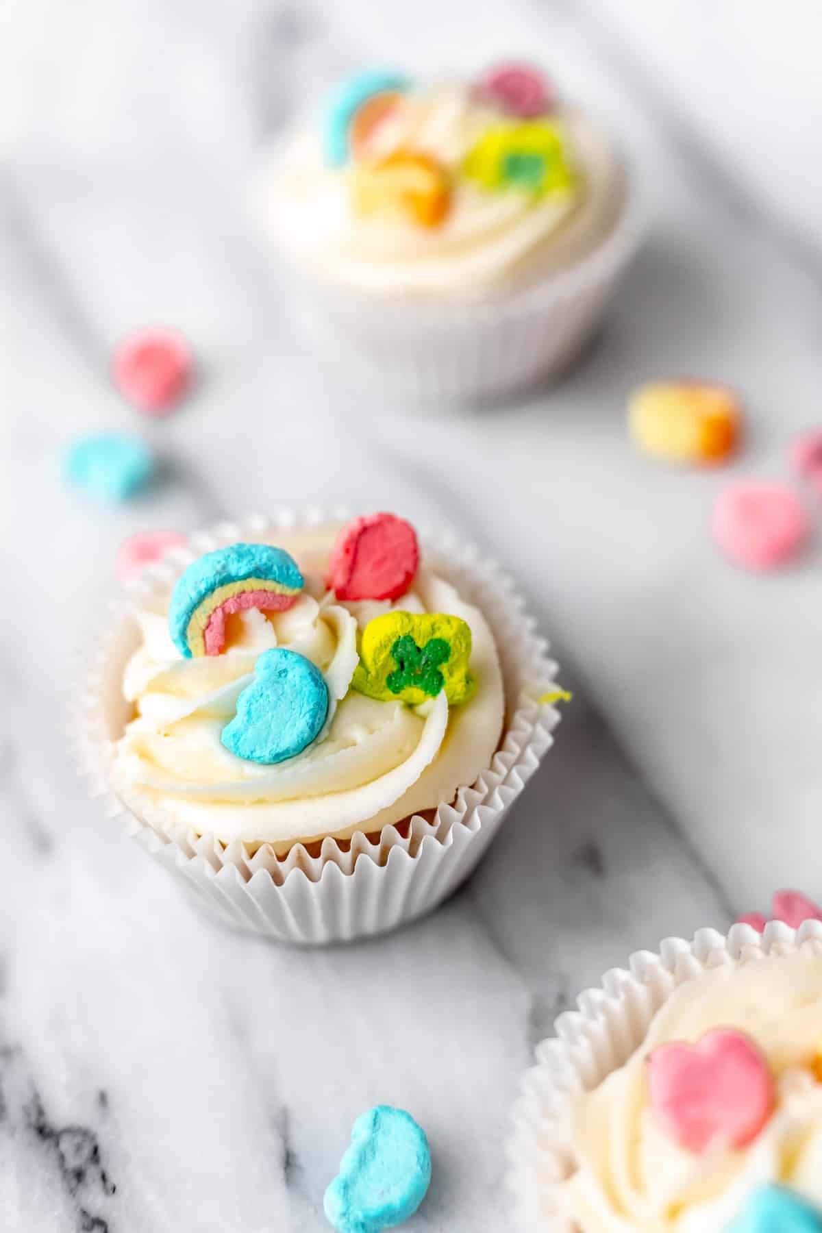 Close up of a Lucky Charms cupcake with other cupcakes and marshmallows around it.
