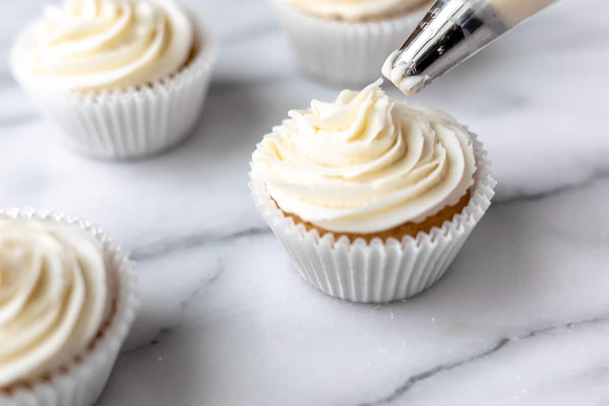 Vanilla frosting being piped onto a cupcake with other cupcakes around it.