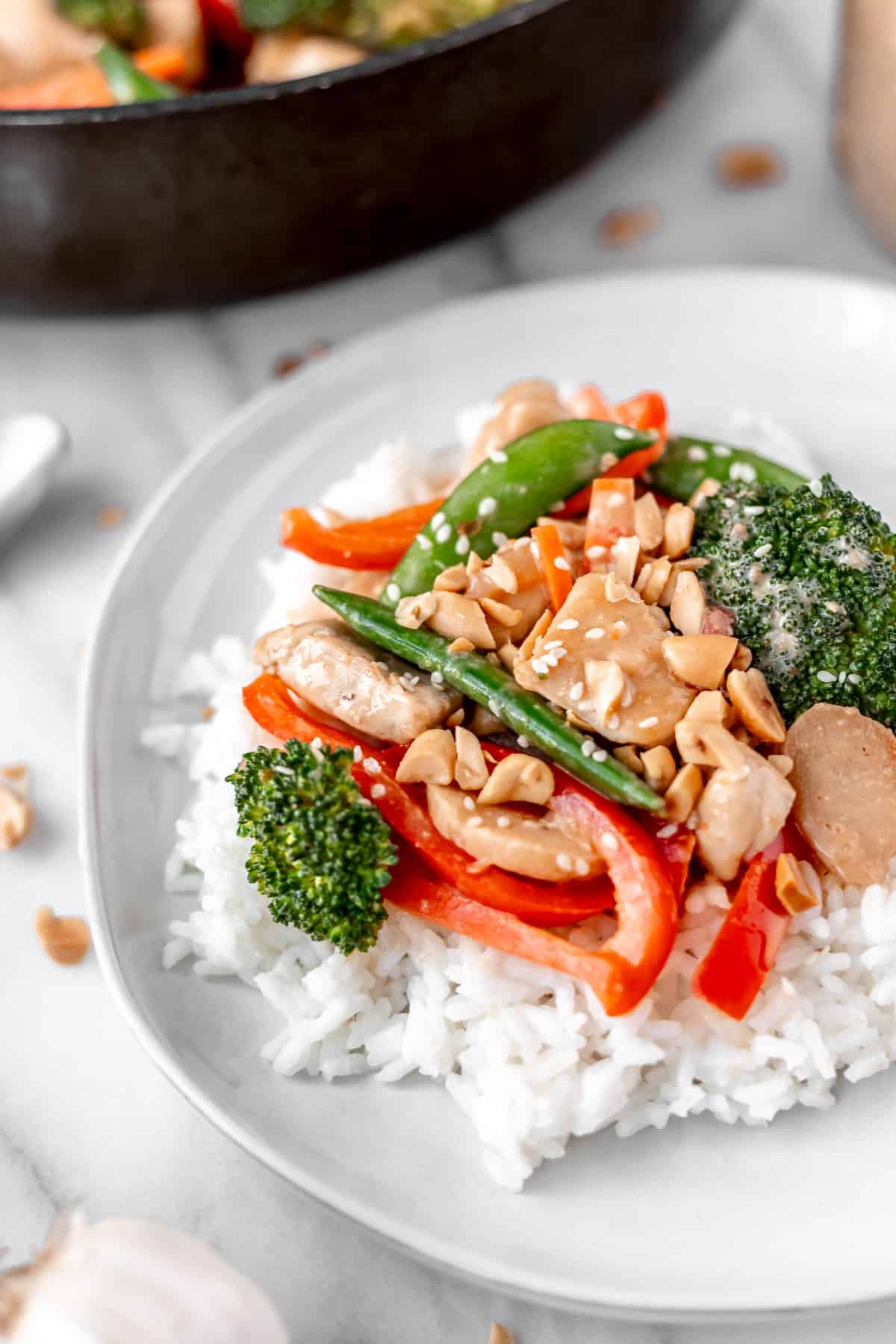 Close up of a serving of chicken peanut stir fry on rice on a white plate with a skillet partially showing in the background.
