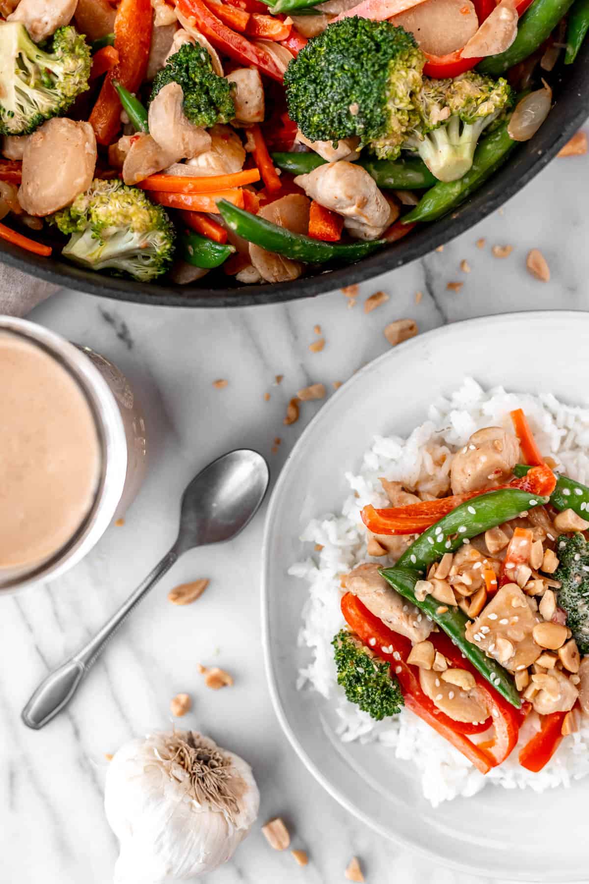Overhead of chicken peanut stir fry in a skillet and on a plate with a jar of peanut sauce, spoon, garlic and peanuts around them.