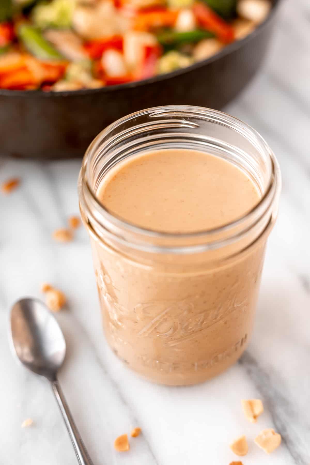 A glass jar of peanut sauce with a spoon next to it and chopped peanut around it.