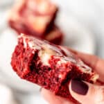 A hand holding up a red velvet cheesecake brownie with a bite taken out and text overlay.