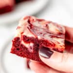 A hand holding up a red velvet cheesecake brownie with text overlay.