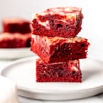 A stack of three red velvet cheesecake brownies on a white plate with a second plate of brownies in the background.