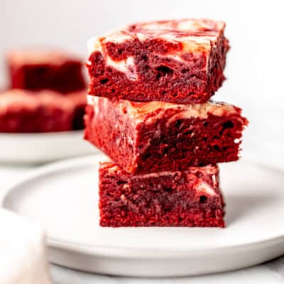 A stack of three red velvet cheesecake brownies on a white plate with a second plate of brownies in the background.