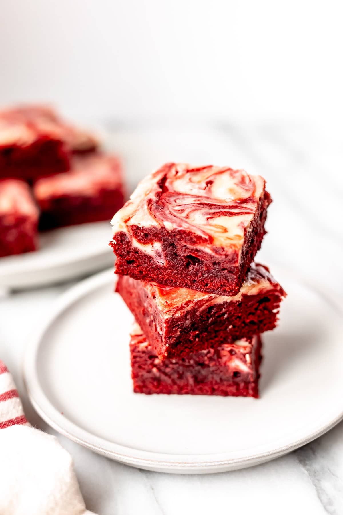 Three red velvet cheesecake swirled brownies stacked on a white plate with more brownies in the background.