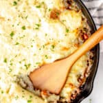 Overhead of a Shepherd's Pie in a cast iron skillet with a wood serving pushing some up with text overlay.