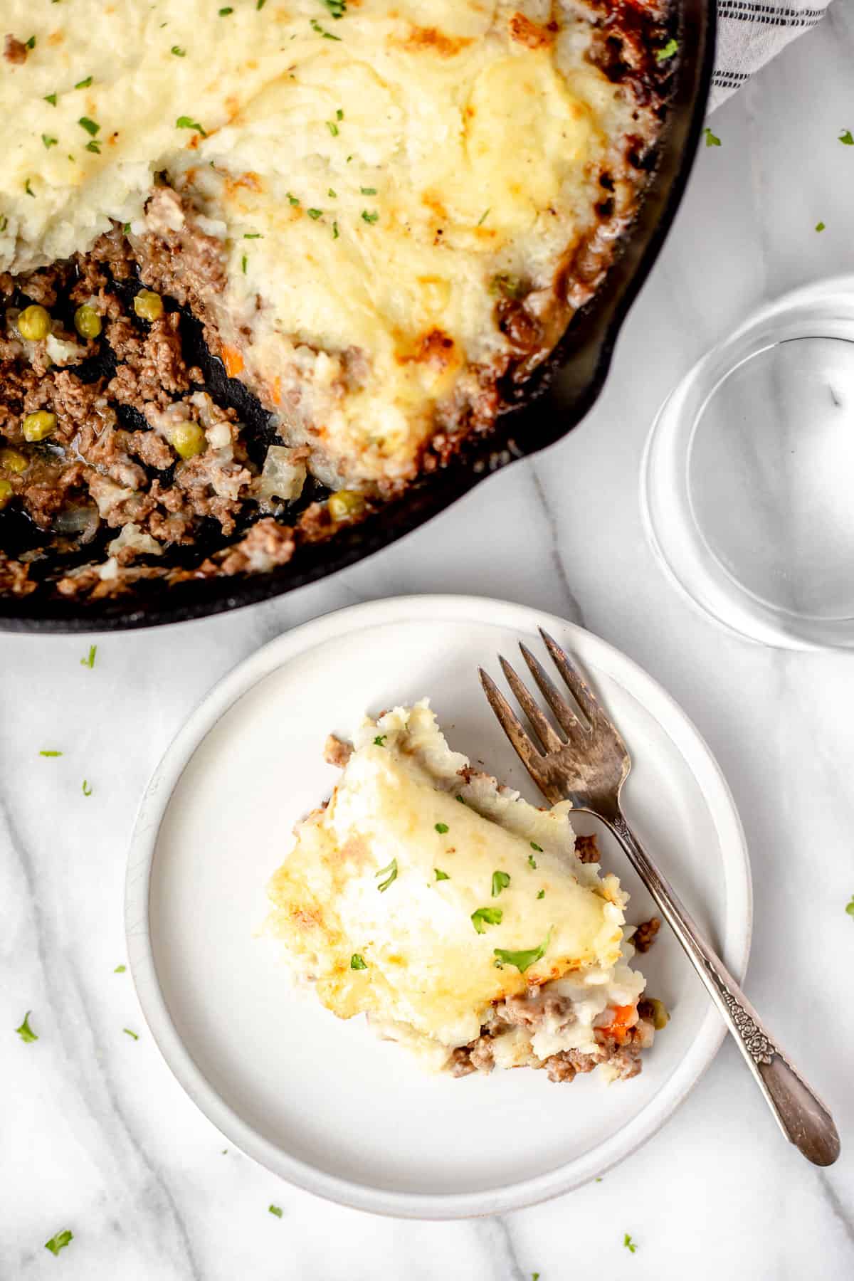 Overhead of a serving of Shepherd's Pie on a white plate with a fork with a cast iron skillet with more Shepherd's Pie partially showing.