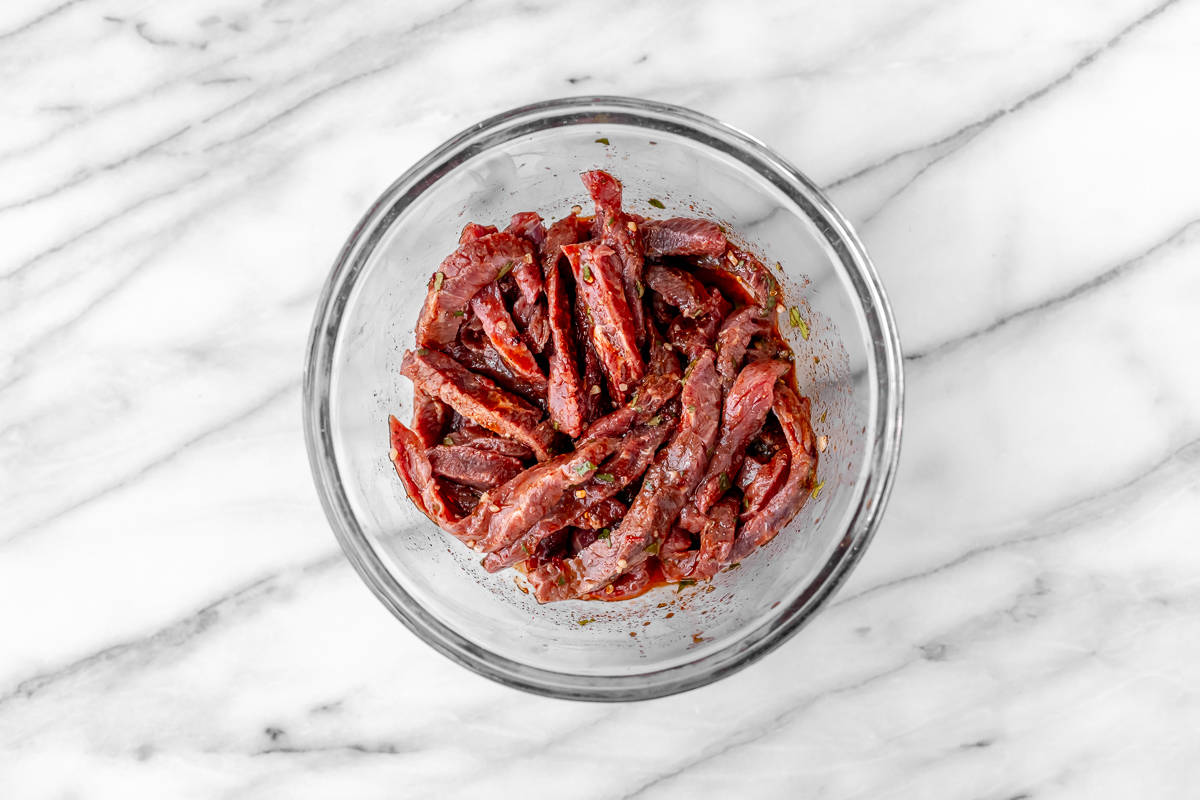 Beef strips in marinade in a glass bowl on a marble background.
