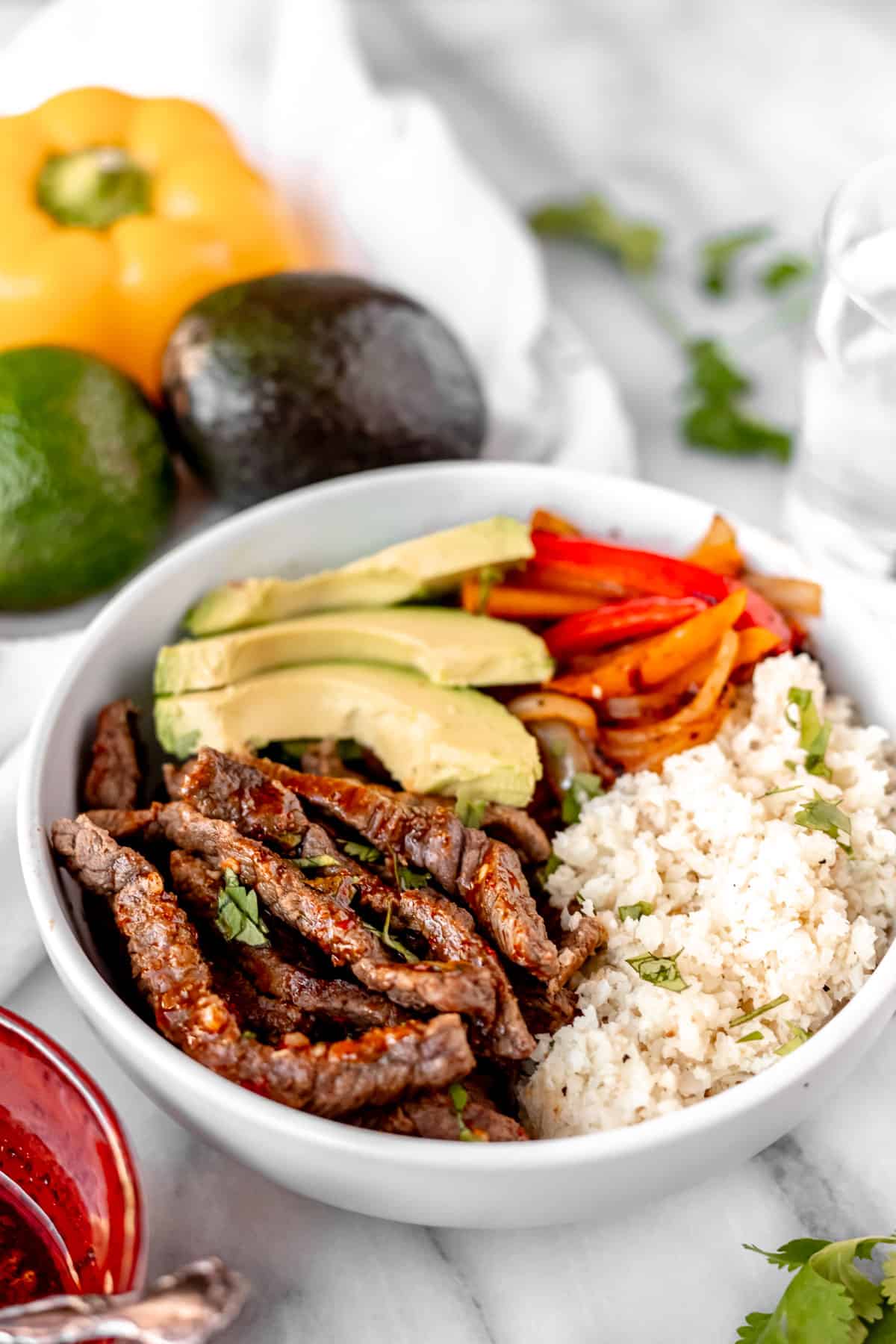 Beef fajita bowl with beef strips, peppers, cauliflower rice and avocado with vegetables and cilantro in the background.