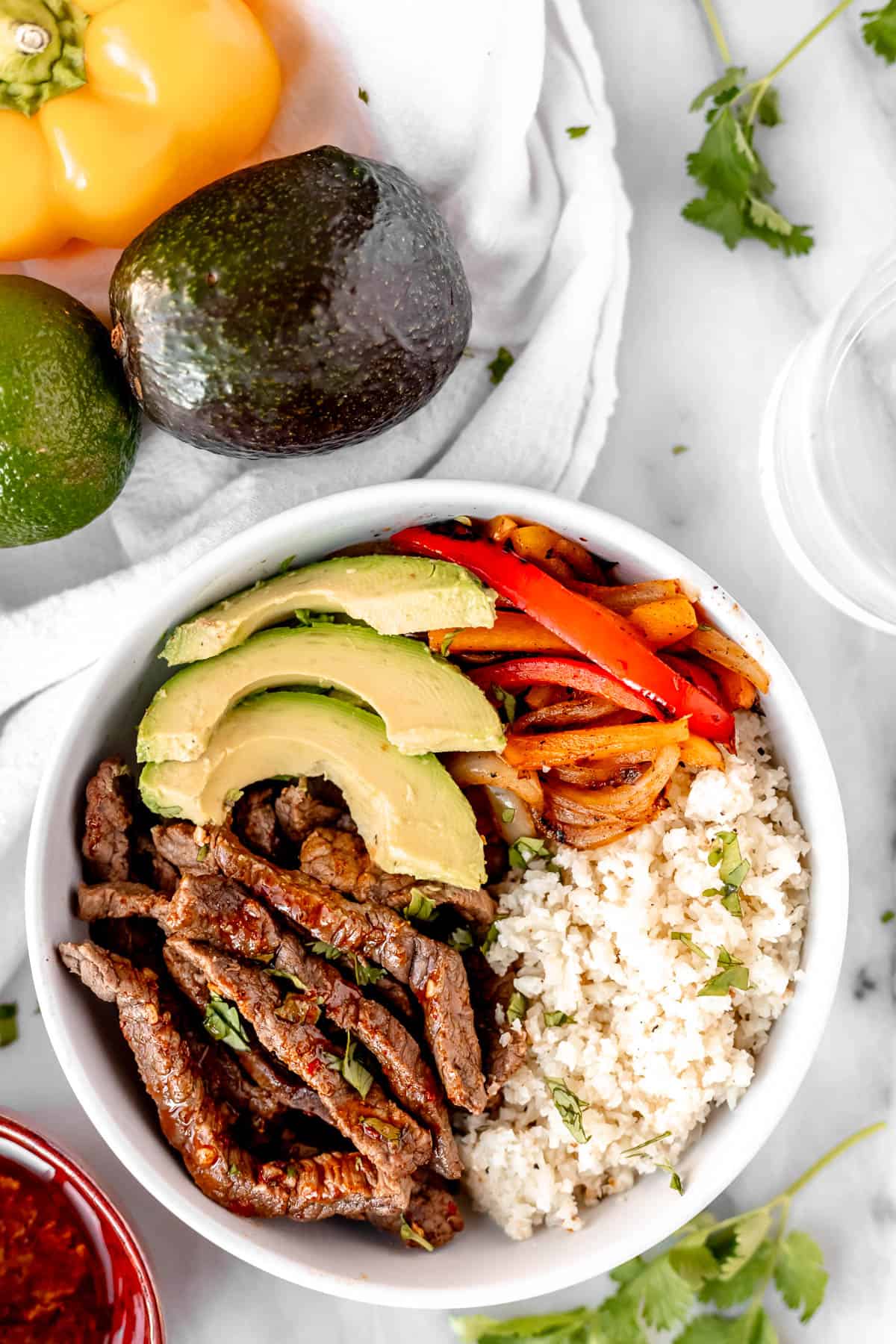 Overhead view of beef fajita bowls with beef strips, peppers, cauliflower rice and avocado with vegetables and cilantro in the background.
