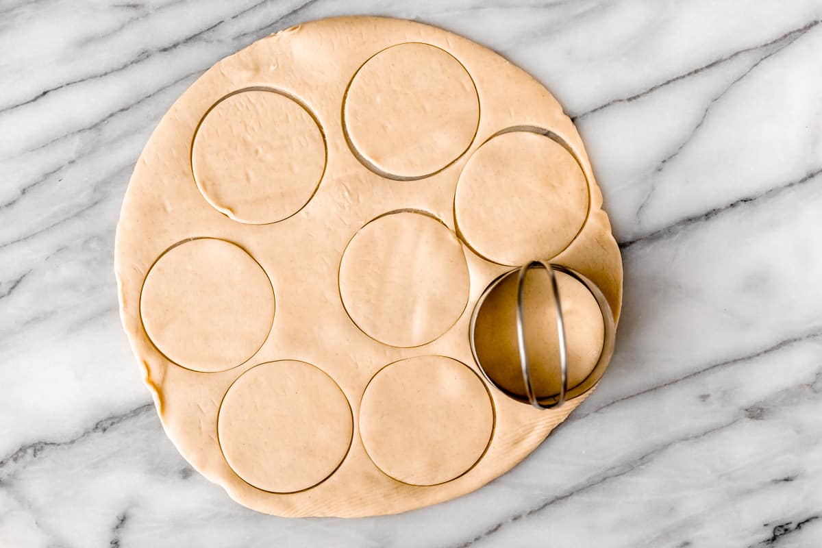 Circles being cit out of a pie crust with a biscuit cutter on a marble pastry slab.