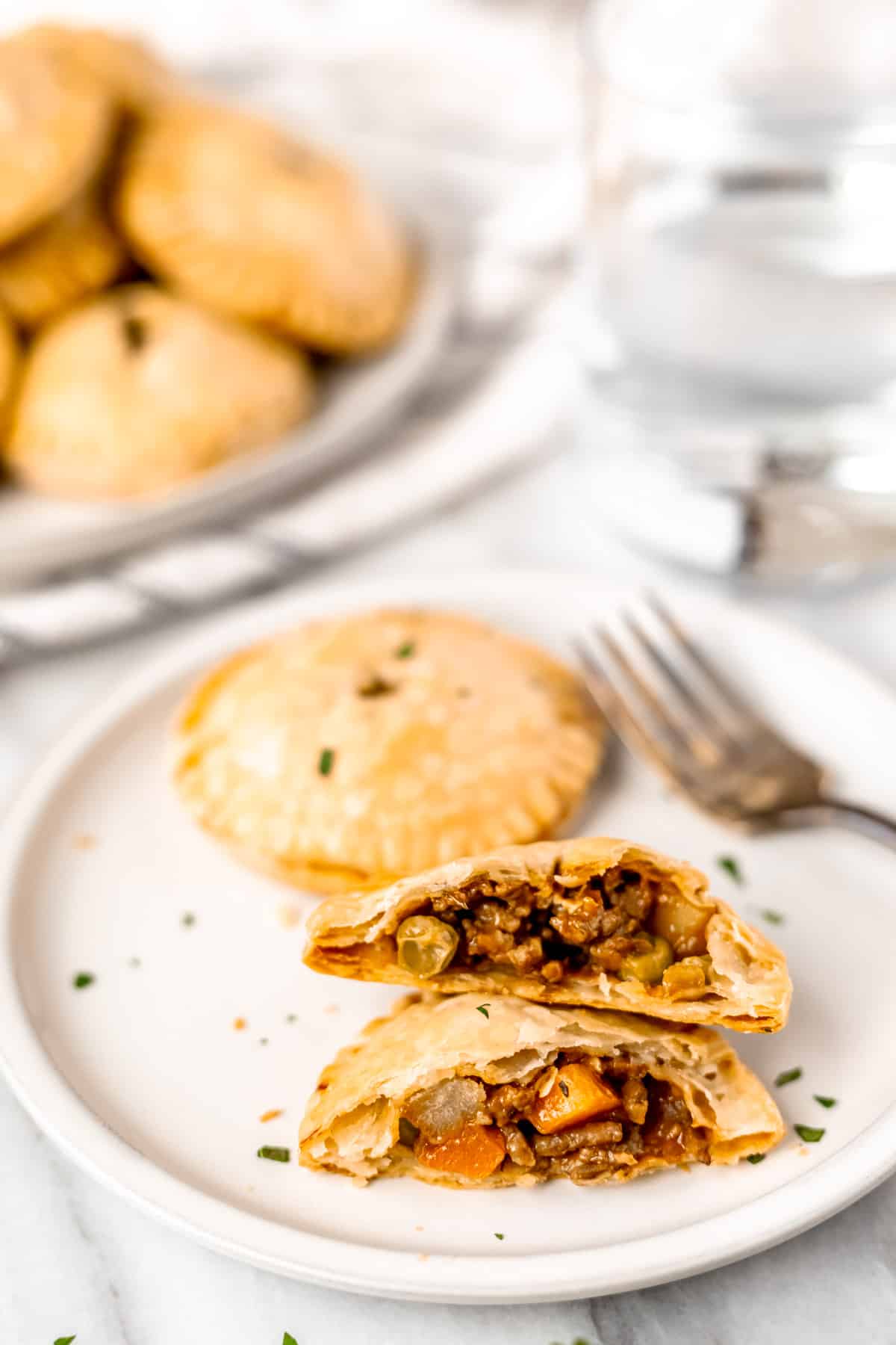 A ground beef hand pie broken in half on a white plate with a fork and second hand pie on it.