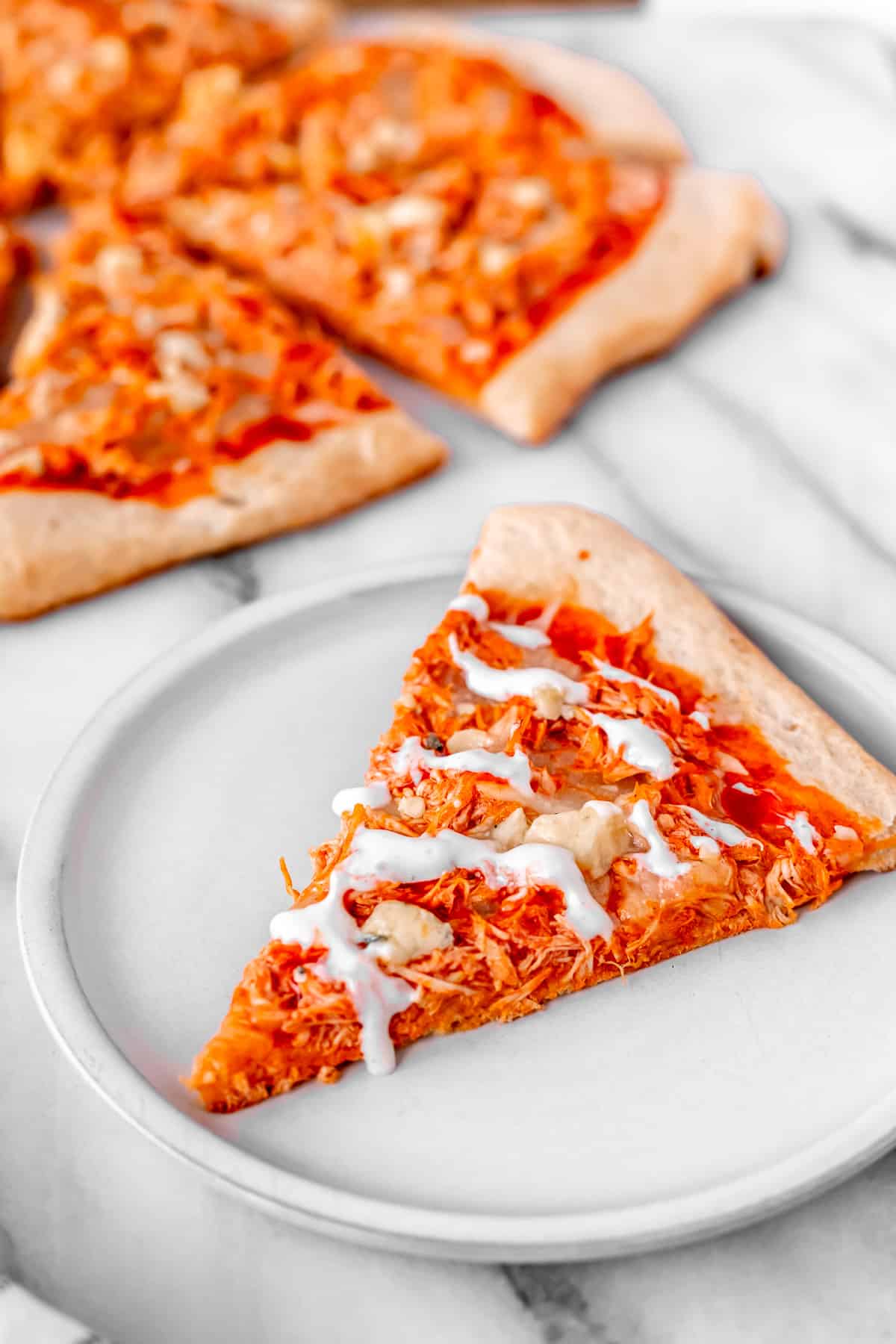 A slice of buffalo chicken pizza drizzle with ranch dressing on a white plate with the rest of the pizza partially showing in the background.