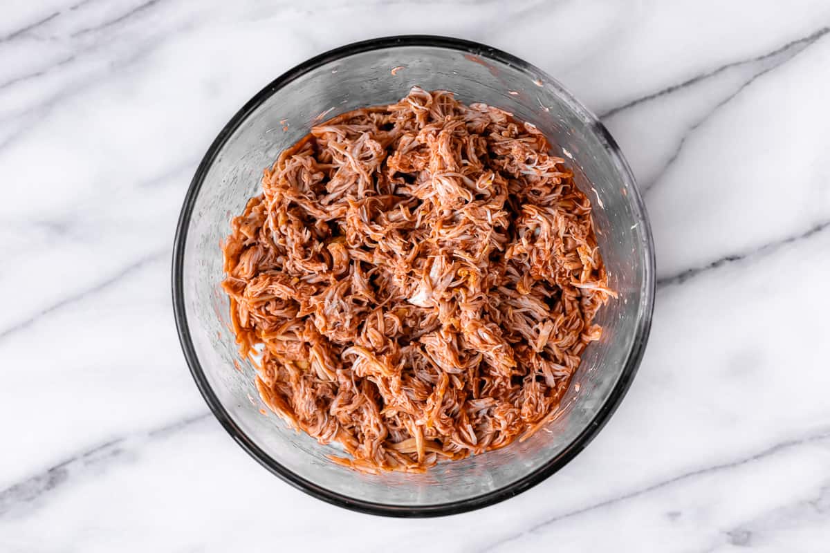 Shredded chicken tossed with buffalo sauce in a glass bowl over a marble background.