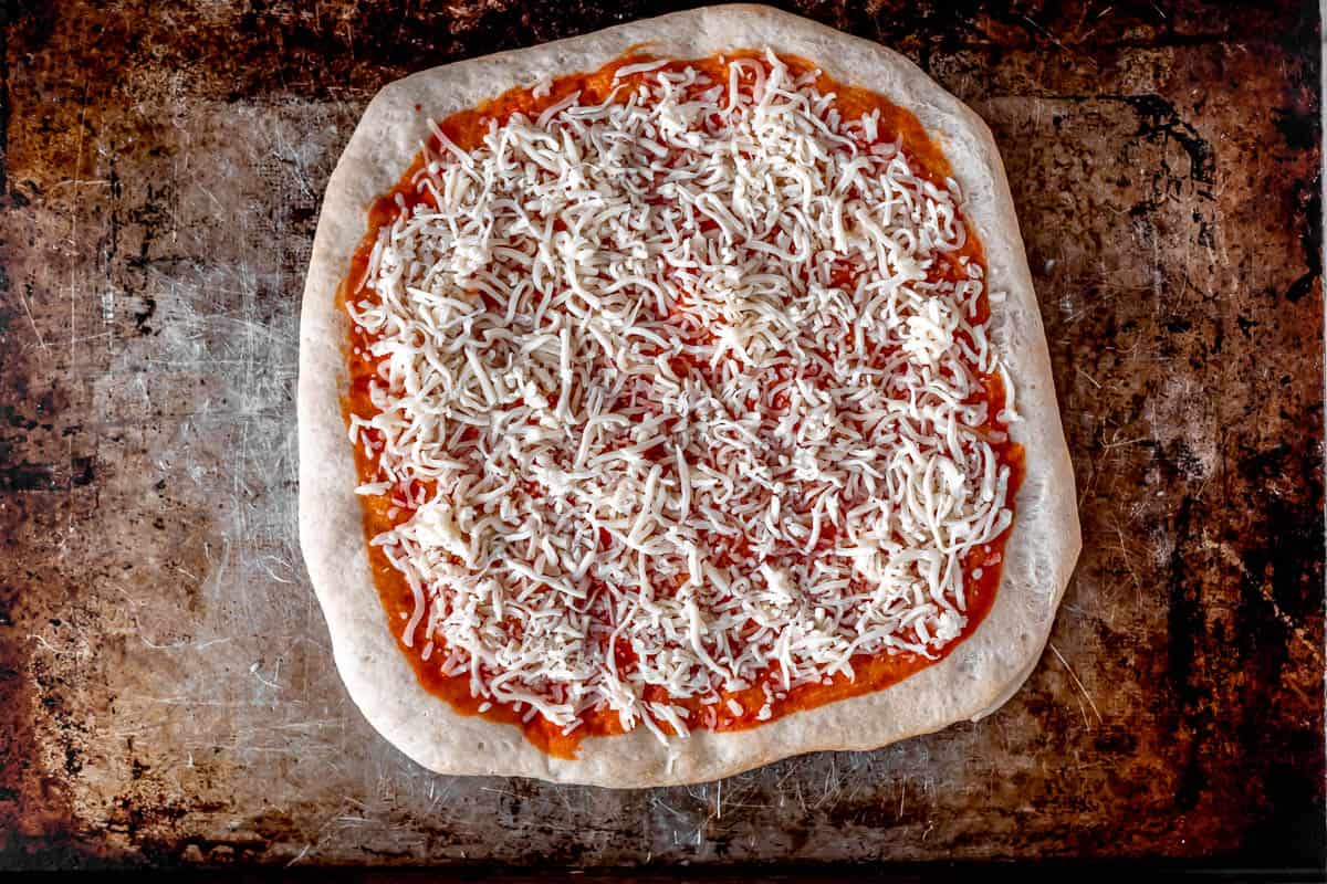 A pizza crust topped with buffalo sauce and shredded mozzarella cheese on a baking sheet.