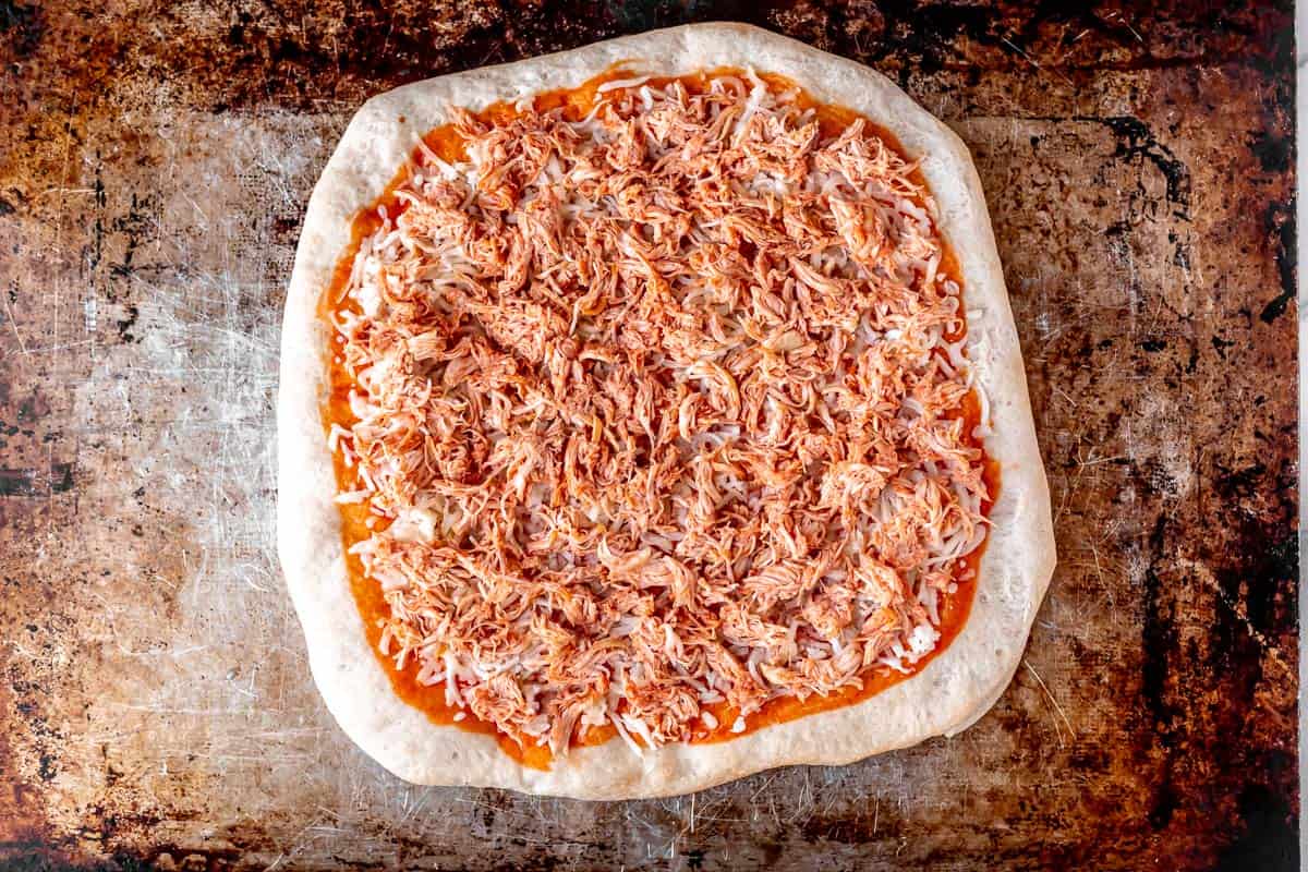 A pizza crust topped with buffalo sauce, mozzarella cheese and shredded chicken on a baking sheet.