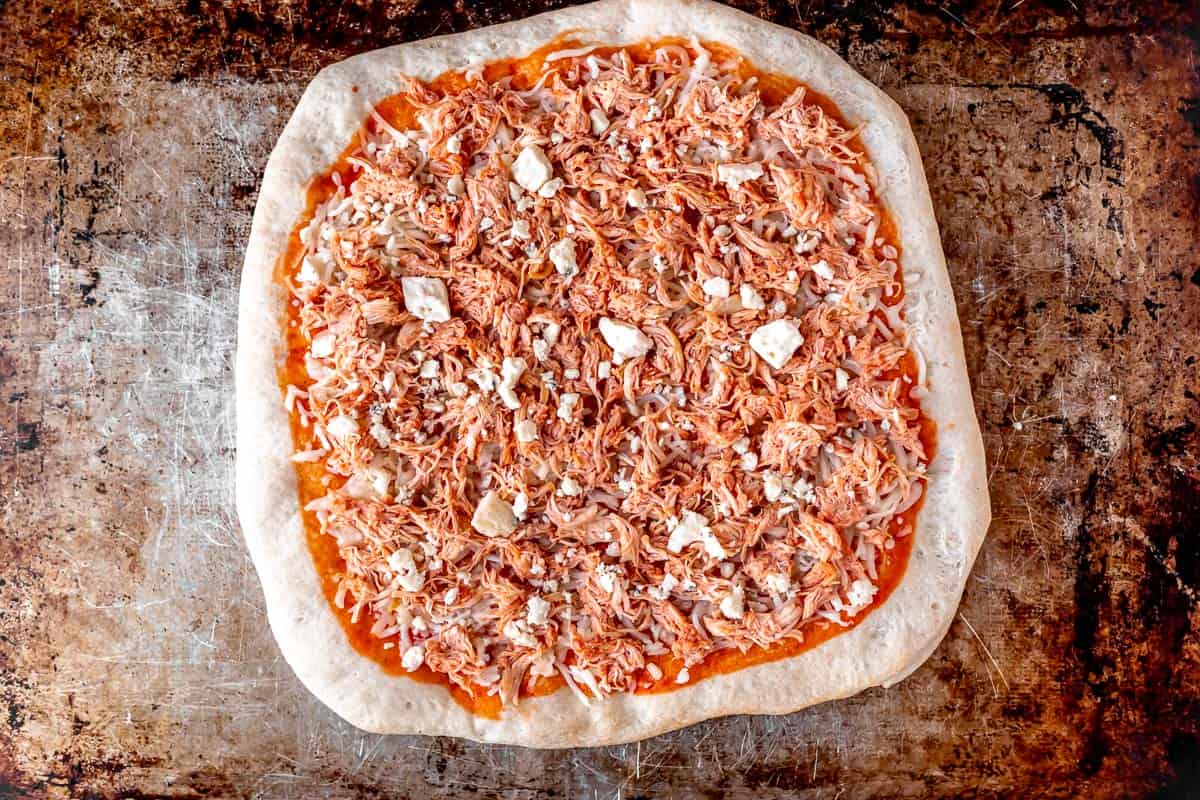 A pizza topped with buffalo sauce, mozzarella and blue cheese and shredded chicken on a baking sheet.