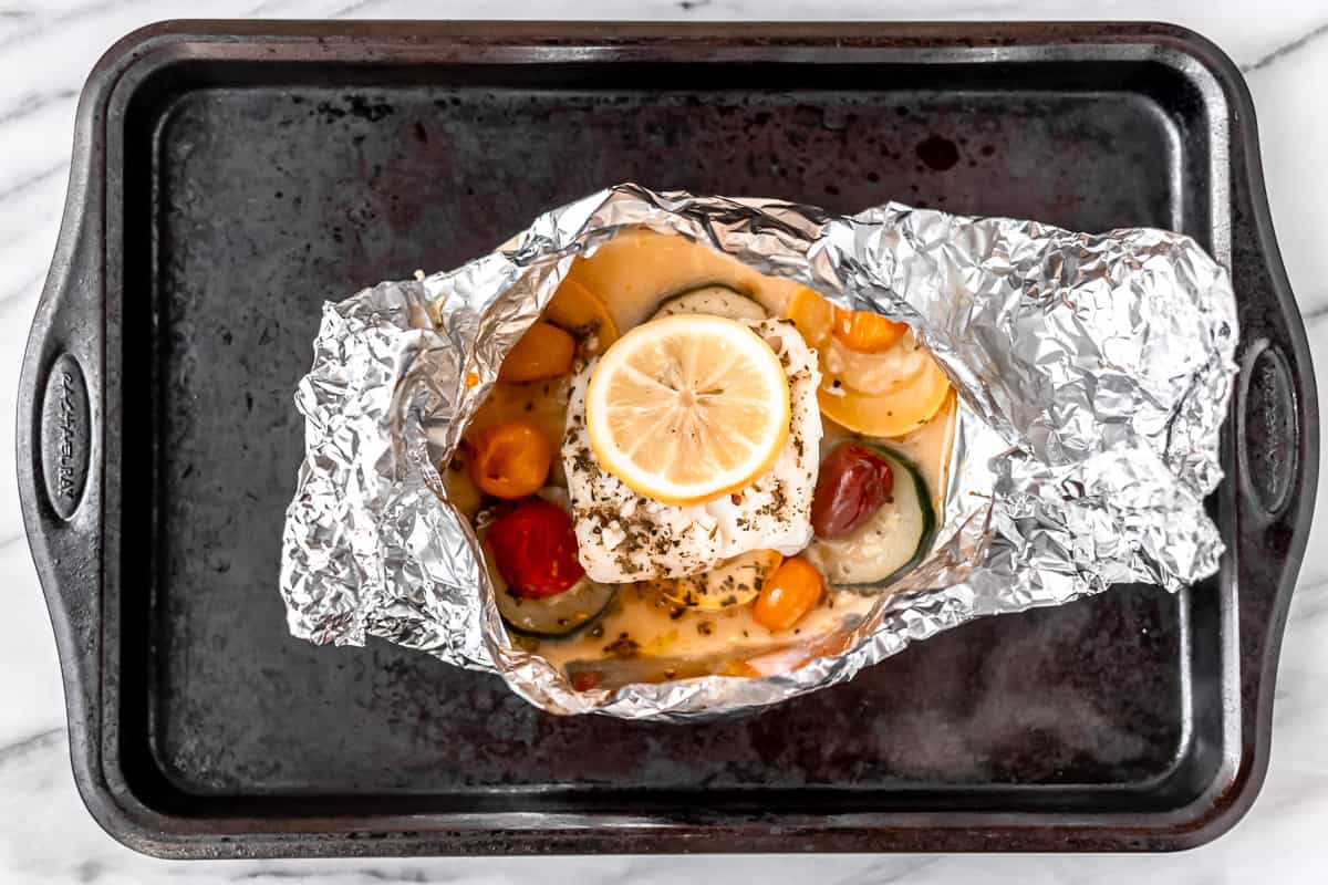 An opened cooked foil packet with cod and vegetables inside on a baking sheet.