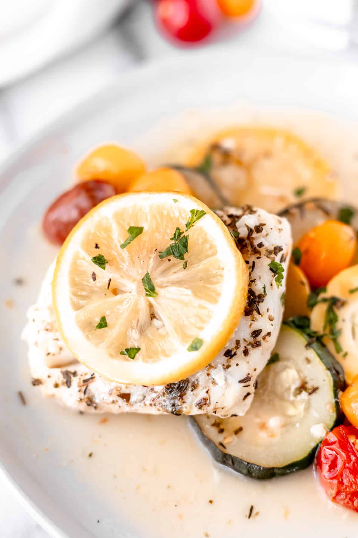 Baked cod topped with lemons on a plate of vegetables such as zucchini and tomatoes in a butter sauce.