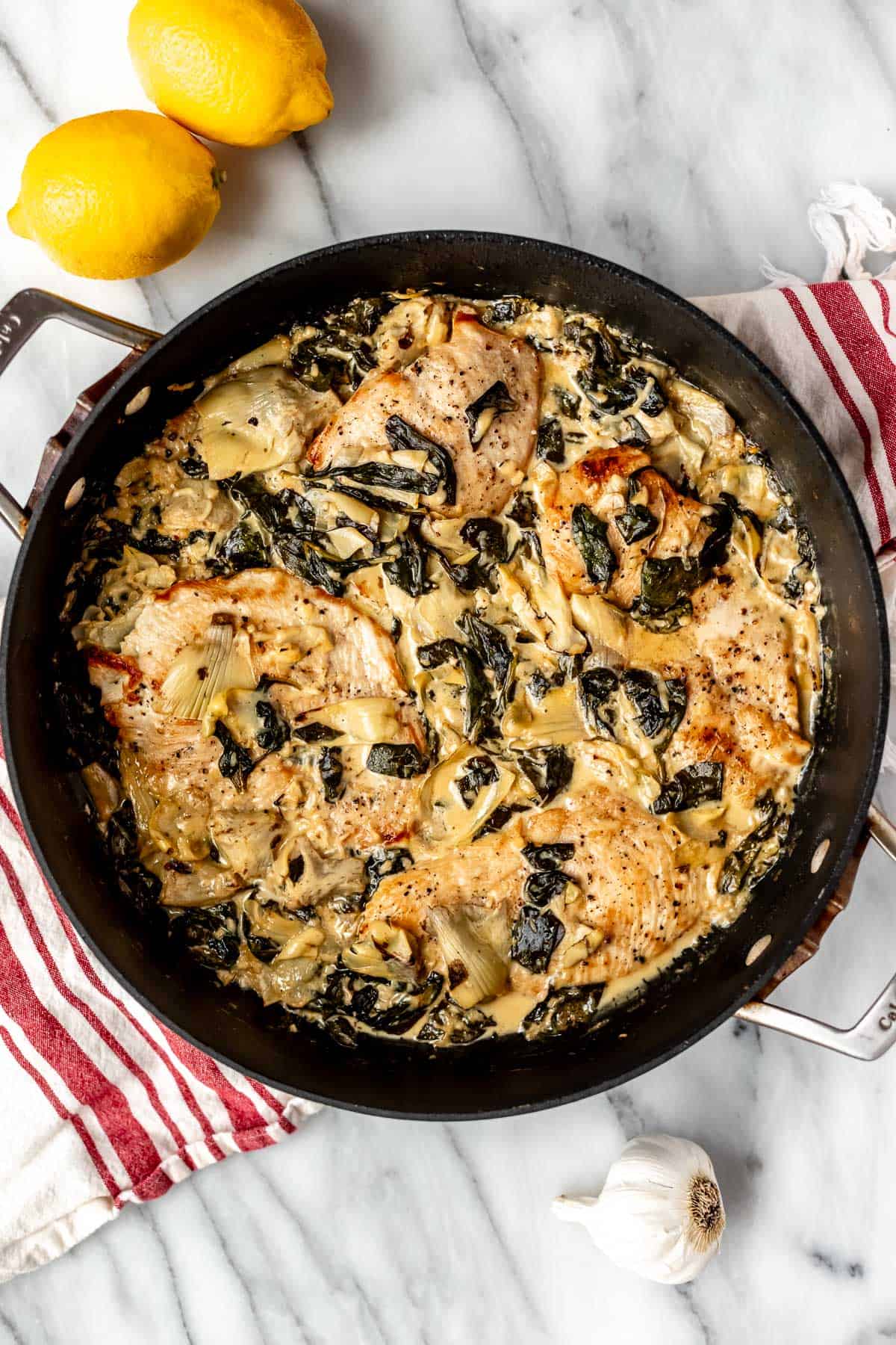 Overhead view of a skillet of spinach artichoke chicken in a skillet on a red and white striped towel with lemons and garlic around it.
