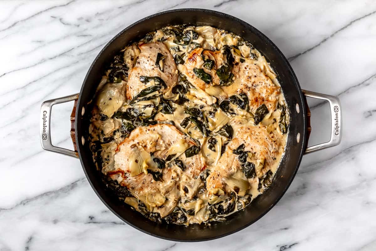 Baked spinach artichoke chicken in a black skillet over a marble background.