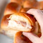 A single ham and cheese slider being held up over the rest of the sliders in a white casserole dish with text overlay.