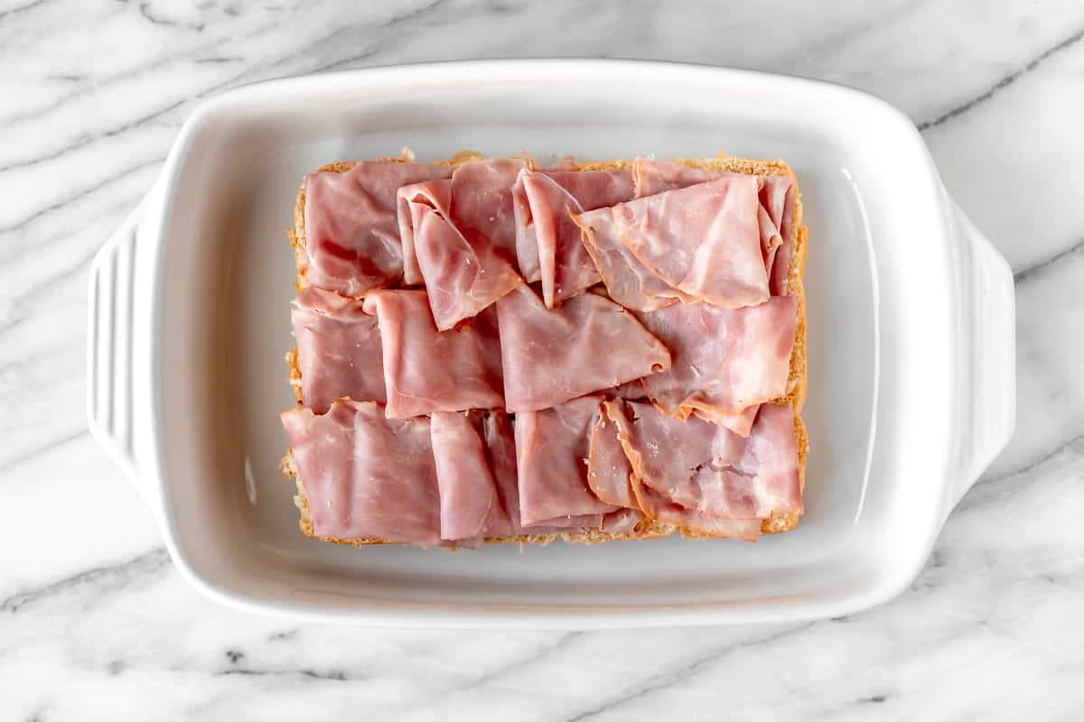 Folded slices of ham on top of the bottom half of rolls in a white casserole dish.