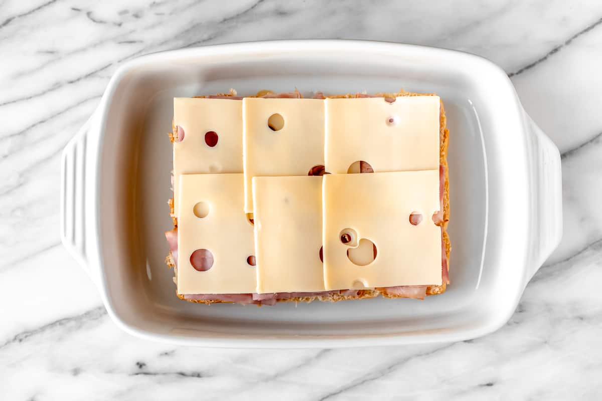 Six slices of Swiss cheese on top of ham and the bottom half of rolls in a white casserole dish.