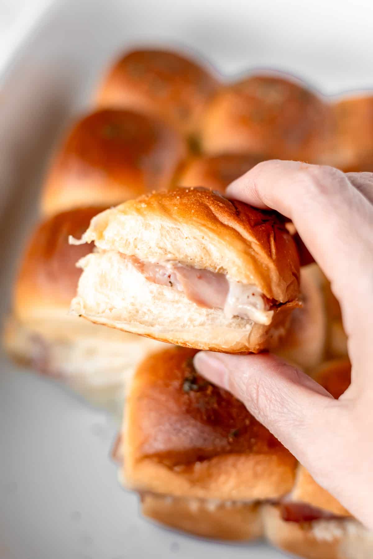 A single ham and cheese slider being held up over the rest of the sliders in a white casserole dish.