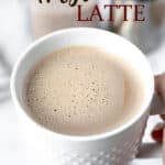 A hand holding up a mug filled with an Irish Latte with creamer and a frothing pitcher in the background with text overlay.