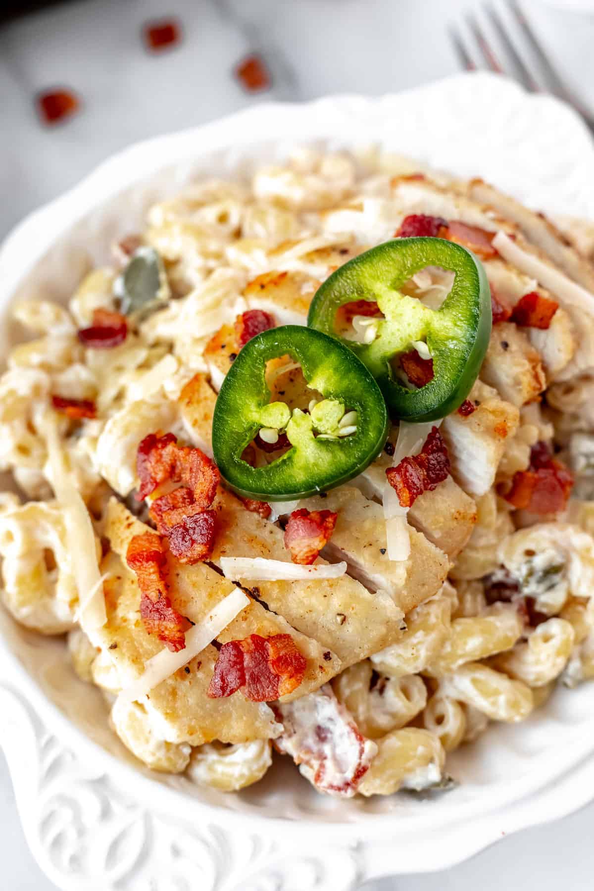 Close up of jalapeno popper chicken pasta in a white bowl with a fork and pieces of bacon in the background.
