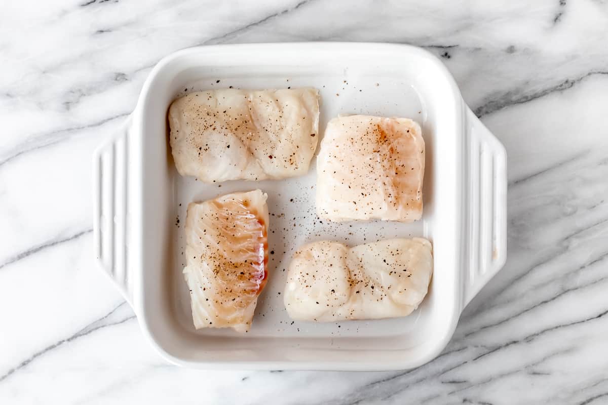 4 cod fillets seasoning with salt and pepper in a white baking dish.