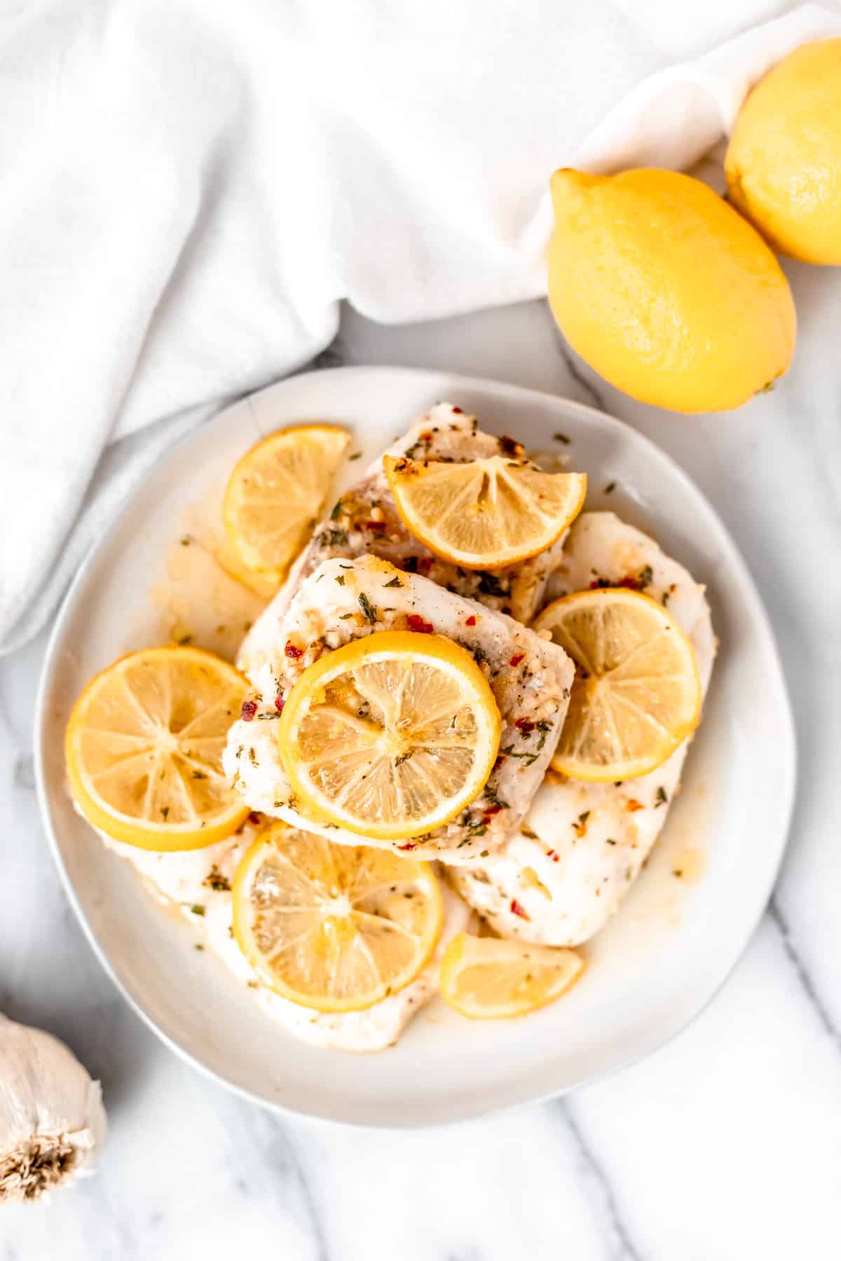Overhead view of lemon butter cod fillets on a white plate with lemon slices on them with a white towel and lemons in the background.