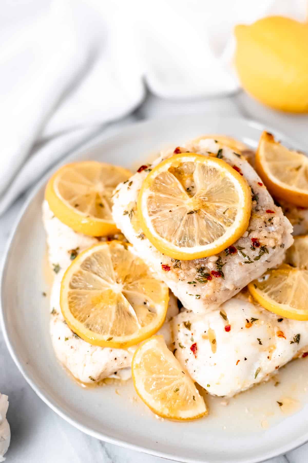 Lemon butter cod fillets stacked on a white plate with lemon slices on and around them.