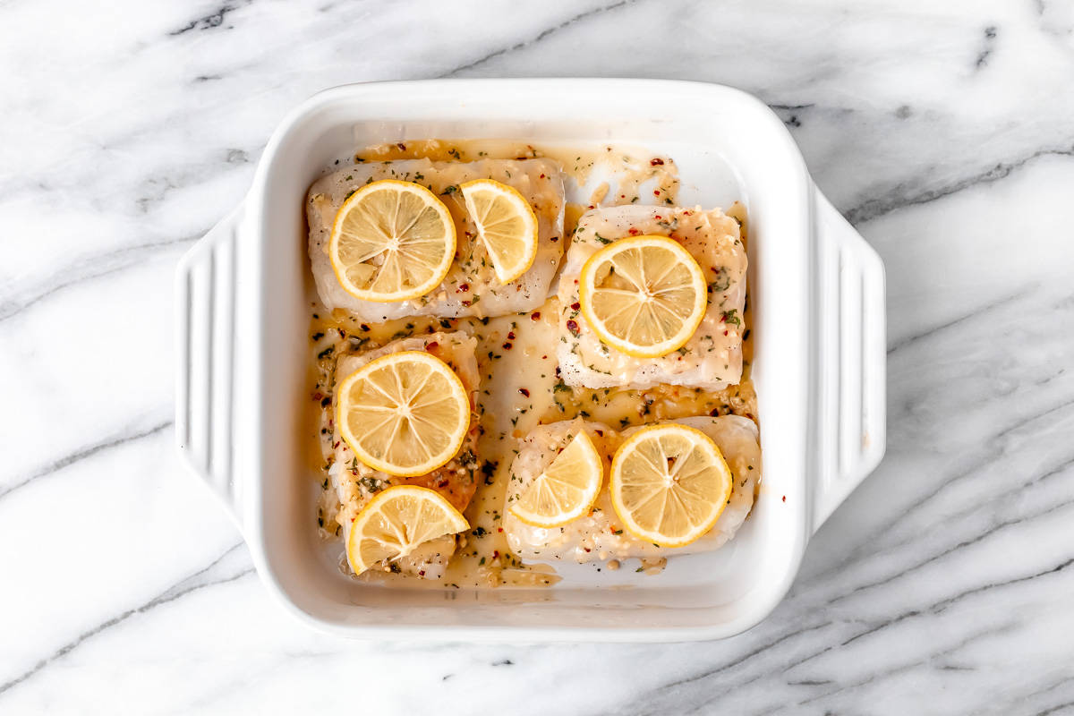 4 cod fillets topped with garlic lemon butter and lemon slices in a white baking dish.