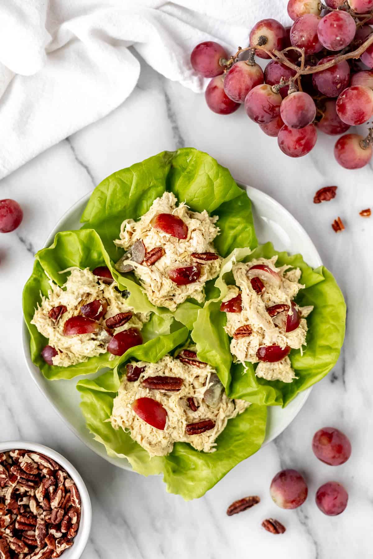 Overhead view of curry chicken salad lettuce wraps on a white plate surrounded by a bowl of pecans, grapes and a white towel.