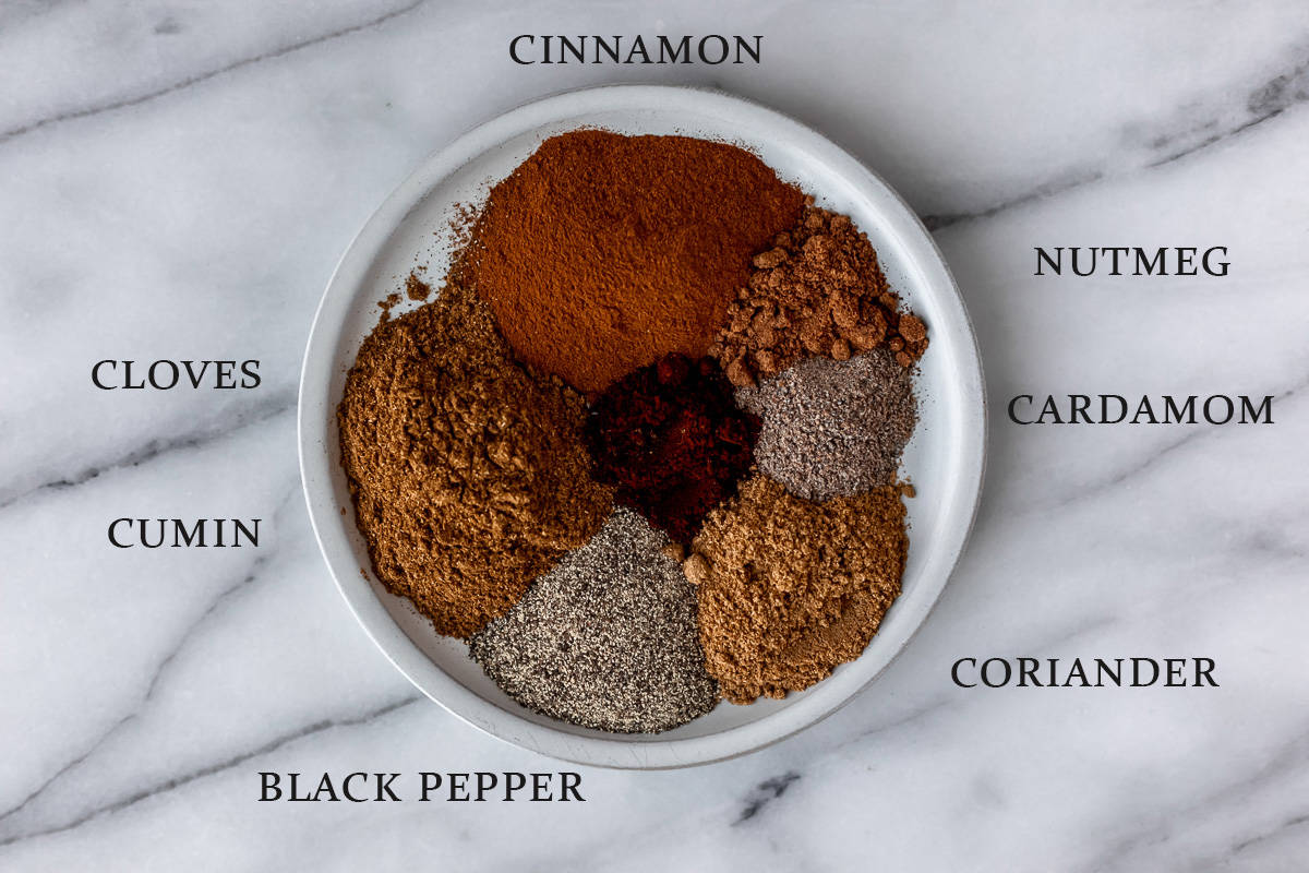 Ingredients needed to make garam masala on a small plate on a marble background with text overlay.