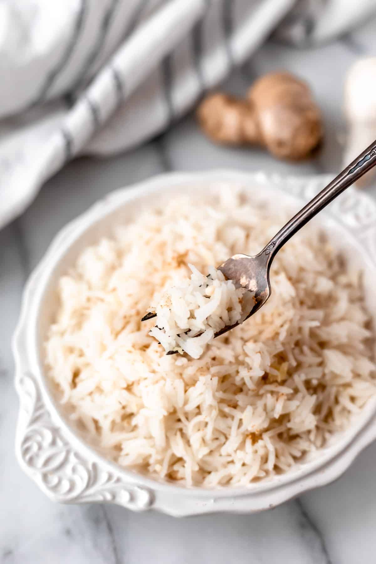 Ginger rice on a fork held up in front of a white bowl of rice with a striped towel and ginger in the background.