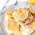 Lemon butter cod fillets stacked on a white plate with lemon slices on and around them with text overlay.