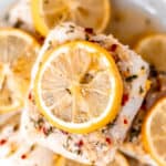 Close up of lemon butter cod fillets stacked on a white plate with lemon slices on and around them.