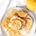 Overhead view of lemon butter cod fillets on a white plate with lemon slices on them with a white towel and lemons in the background with text overlay.