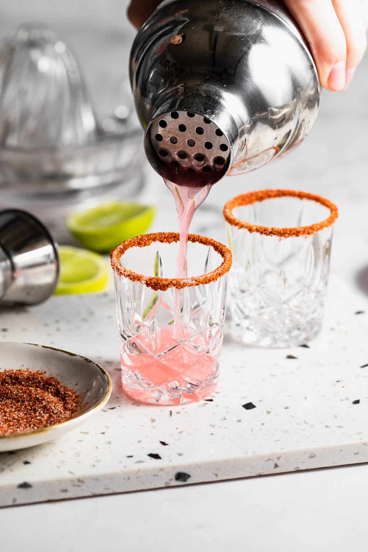A pink liquor being poured into a Tajin rimmed shot glass with a second shot glass and other ingredients around it.