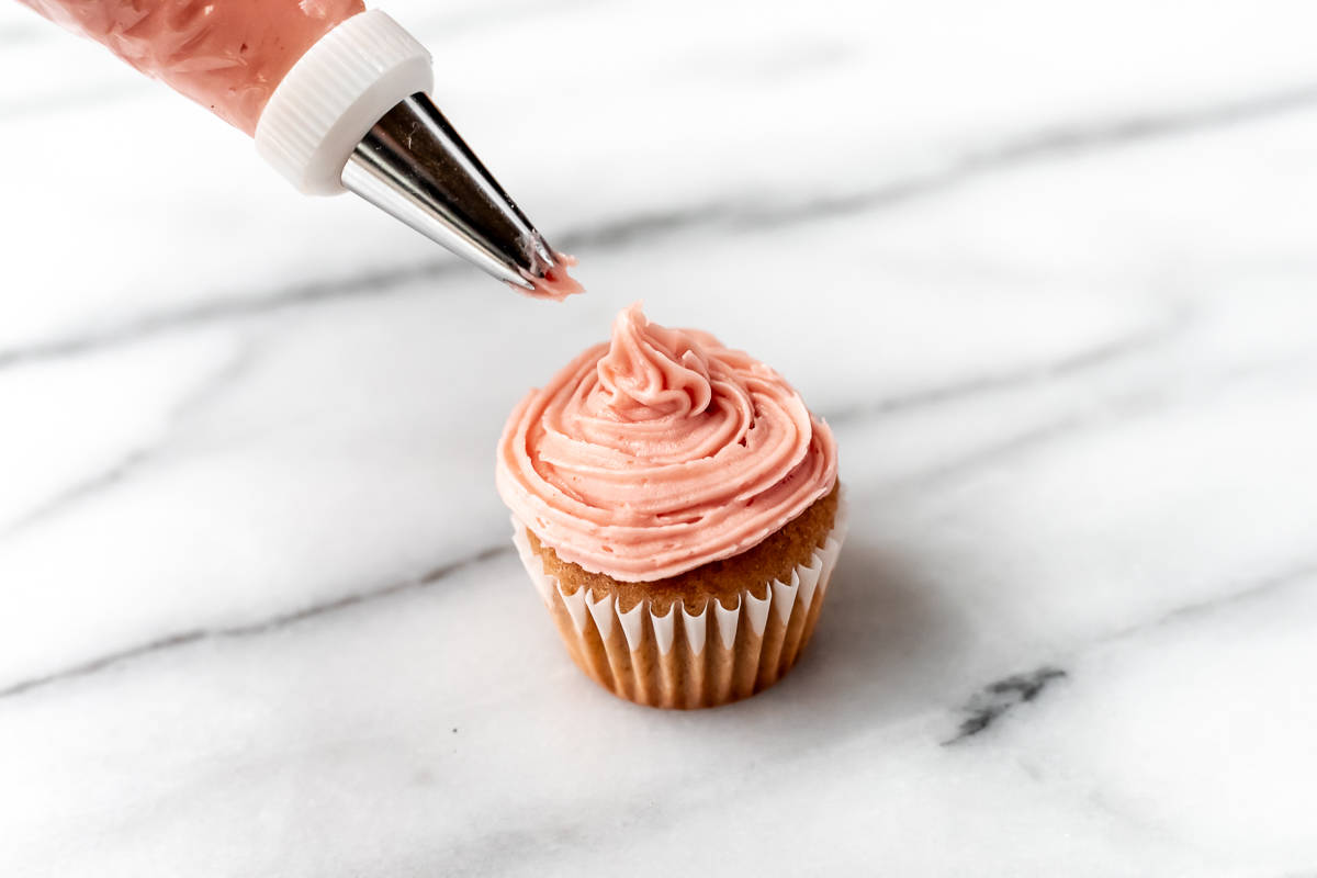 Strawberry frosting being piped onto a mini strawberry cupcake on a marble table.