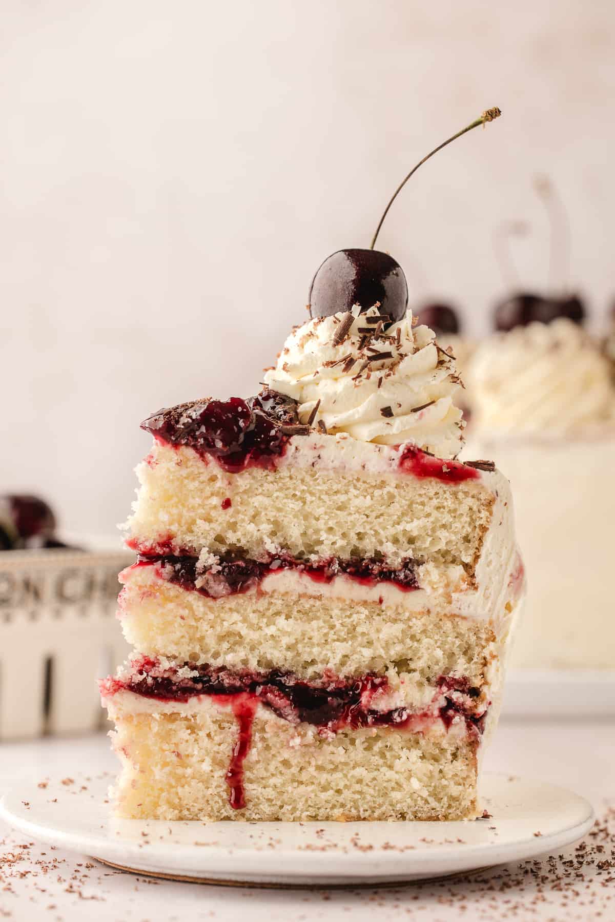 A single slice of white forest cake topped with fresh cherries on a white plate with the remaining cake in the background.