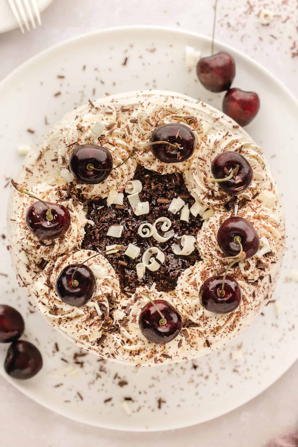 Overhead view of a white forest cake decorated with swirls of frosting, grated chocolate and white chocolate, cherry compote and fresh cherries.